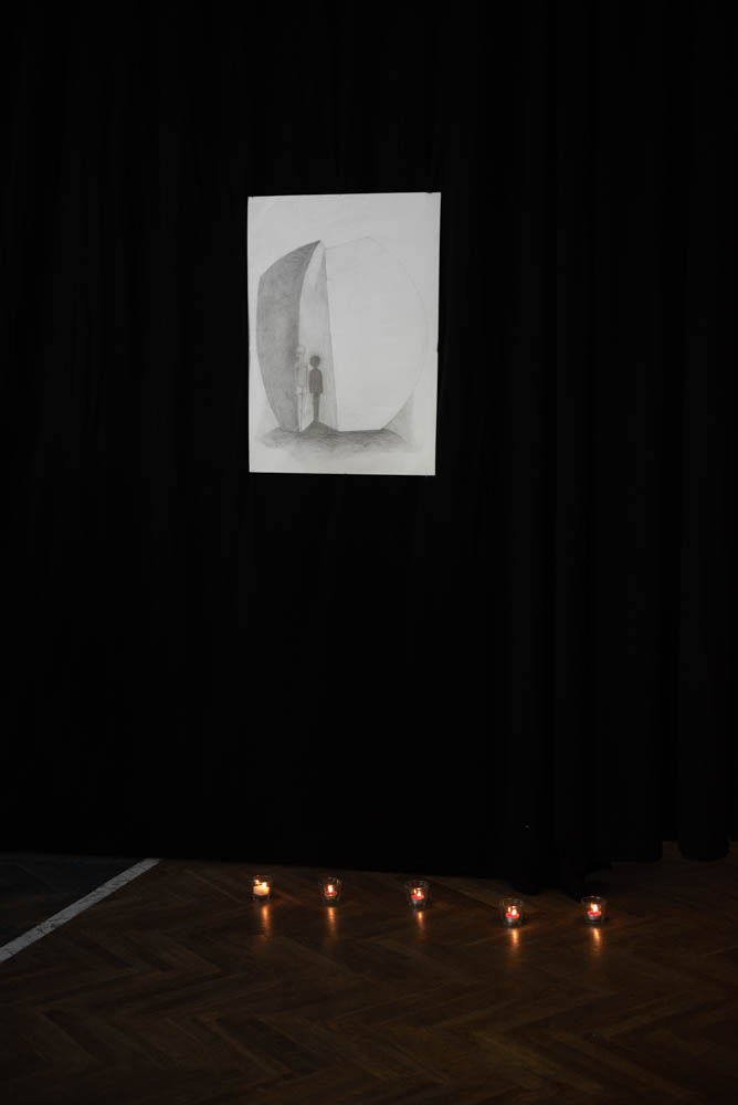 Czarna ściana, a nim plakat przedstawiający Pomnik Pękniętego Serca. Pod plakatem palące się świeczki.